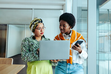 Female coworkers discussing a design brief in office with a laptop and smartphone