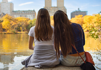 Two girl friends at the city park sitting by a lake 