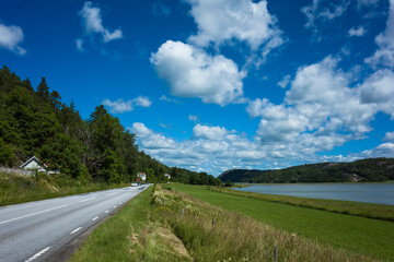 West Coast, Sweden &ndash; A picturesque countryside road runs alongside a serene bay, surrounded by lush greenery and rolling hills. White clouds scatter across the vibrant blue sky on peaceful summer day
