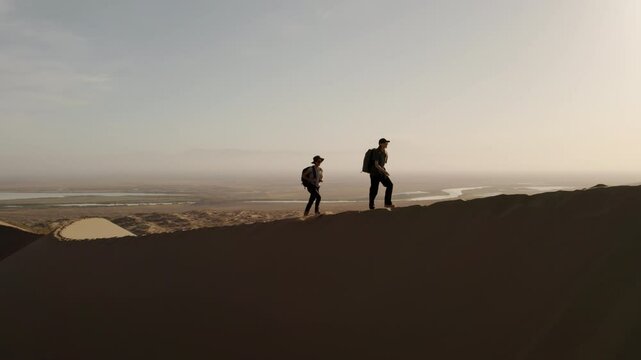 Two young beautiful girls with big backpacks climb a dune above a river valley in the Altyn Emel national park, Kazakhstan