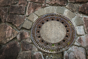 Urban manhole cover surrounded by textured stone wall at a city street during daytime