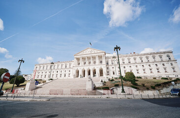 Fototapeta premium A grand neoclassical government building with a wide staircase and columns, set against a blue sky with clouds. The building is surrounded by greenery and street lamps.