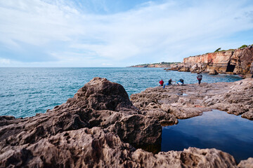 People fishing on rocky cliffs by the ocean under a blue sky.