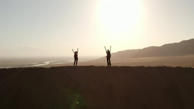 View of two girls standing on top of a high dune and raising their hands towards the sun in the Altyn Emel national park, Kazakhstan