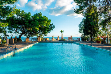 swimming pool with blue water in a hoter resort park during summer tourism season with green trees and blue sea water and cloudy sky on background