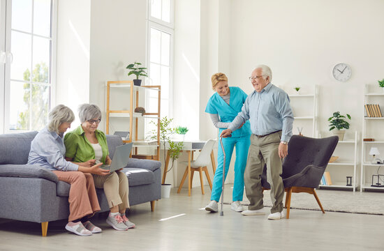 Young friendly female nurse helping a senior patient man to walk with crutches while elderly women using laptop sitting in nursing home. Health care and leisure time in retirement home concept.