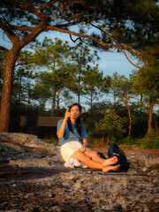 Young woman with pigtails wearing a light blue T-shirt, white shorts, sitting holding a small black camera in front of her, looking at the device with intent, outdoors in a natural environment.