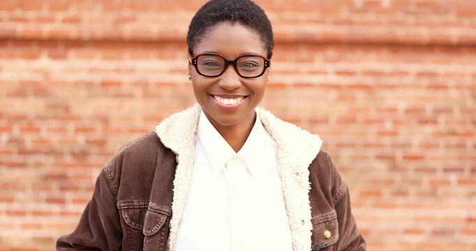 Portrait of a cheerful young black woman wearing glasses and a brown jacket while smiling and posing in front of a brick wall