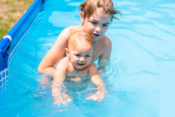Older boy helping toddler stand in swimming pool on sunny summer day