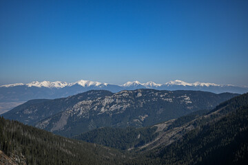 Naklejka premium Stunning panoramic view of a snow-capped mountain range rising above rolling hills and lush forests, creating a breathtaking natural spectacle