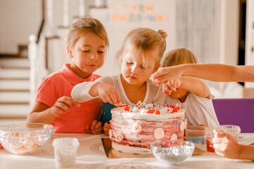 Group of cheerful kids decorating birthday cake together in bright home kitchen during celebration