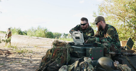 The army officer types reports at a computer in the field. A man with a map joins him, and they review mission details as soldiers in the background secure the camp.
