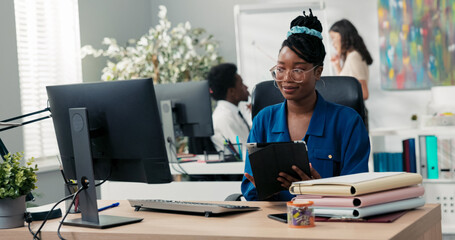 A stylish corporate employee sits in a comfortable chair at her workstation, holding a tablet,...