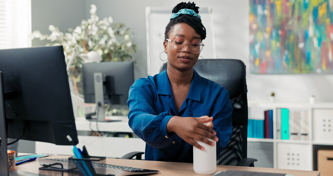 An attractive woman in a shirt and glasses comes to work, sits down at desk, disinfects her hands with a liquid before clicking fingers on the computer keyboard, company health and safety - Powered by Adobe
