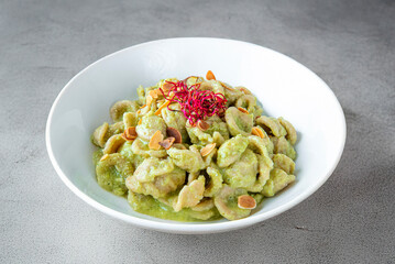 A plate of orecchiette with broccoli pesto in a white bowl, garnished with toasted almonds and pink microgreens. The green sauce contrasts with the gray countertop, offering a modern, fresh look.