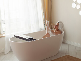 Cheerful young Caucasian beautiful female lying in bathtub with foam in good mood smiling and...