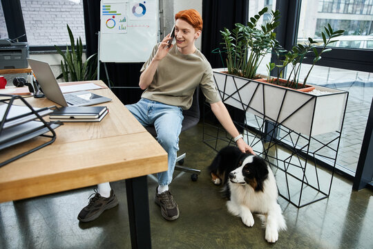 Focused young man with red hair enjoys work while interacting with pet dog in modern office