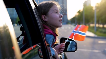 Beautiful small teen looking out of a car window holding Norwegian flag in hands traveling in...