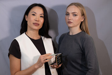 A professional makeup artist and a model pose together in a studio while holding beauty products. They are dressed stylishly, with a neutral background emphasizing elegance, professionalism