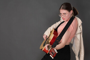 Female guitarist strumming her Spanish six string acoustic guitar and looking directly at camera. 18 year old woman guitar player dressed pink t-shirt and beige leather jacket thrown over one shoulder