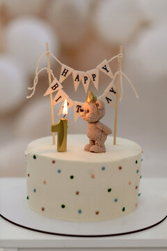 Minimalist first birthday cake with a teddy bear figurine, a lit number one candle, and a 'Happy Birthday' banner. Soft pastel tones and festive decorations create a warm, celebratory atmosphere.