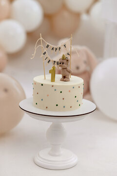 Minimalist first birthday cake with a teddy bear figurine, a lit number one candle, and a 'Happy Birthday' banner. Soft pastel tones and festive decorations create a warm, celebratory atmosphere.