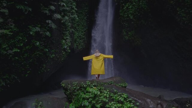 Blonde girl at waterfall flowing in Bali raises hands up. Hiking, Woman in a yellow raincoat or jacket in the tropical jungle enjoys a vacation or trip, lifts palms. Rainy season, Indonesia.