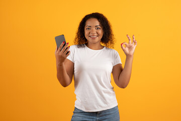 Young African American woman smiling, holding smartphone in one hand and showing OK sign with...