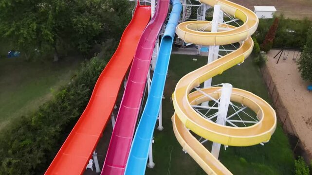 Drone flying backwards over a large water slide split into four sections: orange, red, blue, and yellow, with a rider on the blue slide. Surrounded by grass and trees in a water park.