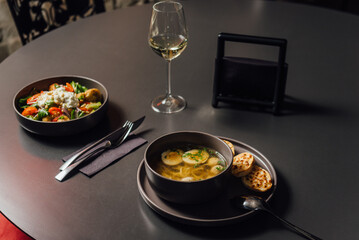 Chicken soup with egg noodles, spinach, broccoli and parmesan croutons, alongside a Panzanella with stracciatella – a Tuscan summer salad. Minimalistic dining scene with a glass of wine, copy space