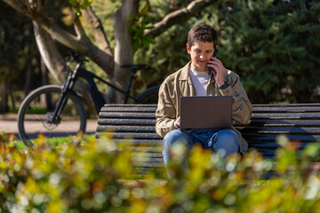 Chico con ordenador negro hablando con telefono movil en un banco en el parque un dia soleado con...