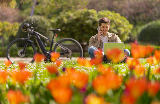 Joven caucasico hablando con telefono movil en el parque sentando en el cesped con portatil metalico y bici electrica de fondo