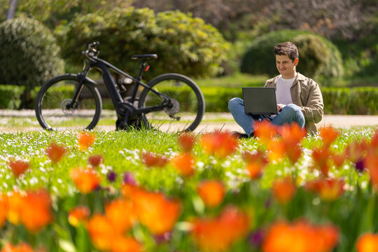 Joven caucasico sonriente en el parque sentando en el cesped con portatil negro y bici electrica de fondo