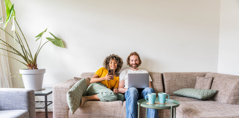 Multiracial young couple using computer laptop and smart mobile phone sitting on the sofa at home - Happy diverse husband and wife using pc online services - Technology life style concept