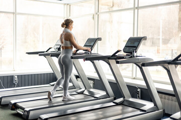 Happy European woman in sportswear and wireless headphones and towel around neck doing hard cardio workout on treadmill, lady working out in empty gym