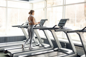 Fototapeta premium Happy European woman in sportswear and wireless headphones and towel around neck doing hard cardio workout on treadmill, lady working out in empty gym
