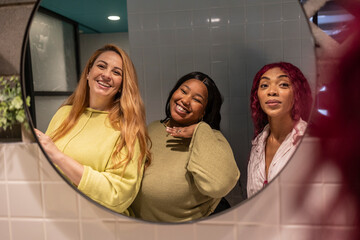 Three happy female colleagues smiling in the office bathroom mirror