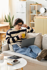 Young man engaging in online studies while relaxing at home with coffee in hand