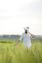 A Pakistani Indian farmer in traditional white attire walks through a green wheat field, carrying a pickaxe on his shoulder while touching the crops.