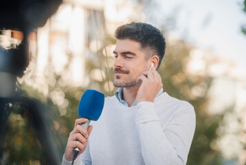 Male journalist wearing earpiece is reporting live using microphone outdoors
