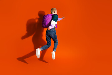 Fototapeta na wymiar Vibrant young student jumping energetically with backpack against bright orange background, dressed in trendy casual attire