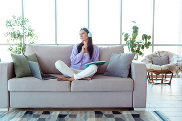 Fototapeta premium Brunette woman sitting on a sofa in a cozy living room environment wearing headphones and holding a book with relaxed thoughtful expression