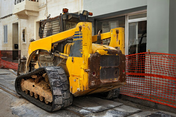 An old yellow mini track loader at a city construction site. Rusty, scratched metal, tracks covered in mud against a building. The construction area is protected by a red mesh fence.