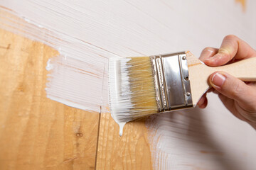 Close-up of a hand holding a paintbrush dripping with white paint, brushing a wooden wall.