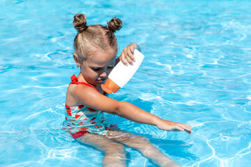 Cute little girl sitting in hotel swimming pool and applying sunscreen to take care of her skin. Summer vacation concept. advertising of tour operators and all-inclusive hotels