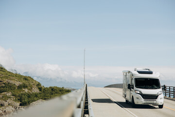 A modern white motorhome drives along a scenic mountain road with a breathtaking view of distant misty peaks and lush green hills under a clear blue sky. 