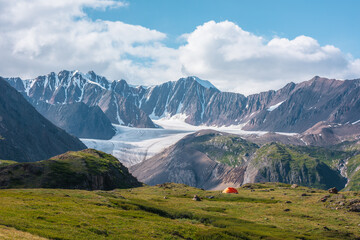 Colorful landscape with orange tent in alpine valley with view to big glacier tongue and large snow-capped mountain range under clouds in blue sky. Green hills and rocks against ice and sheer crags.
