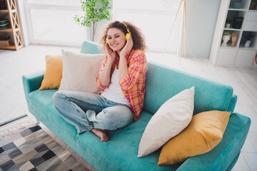 Fototapeta premium Young woman with curly hair sitting on a cozy sofa in a bright living room listening to music and enjoying a relaxing weekend indoors.