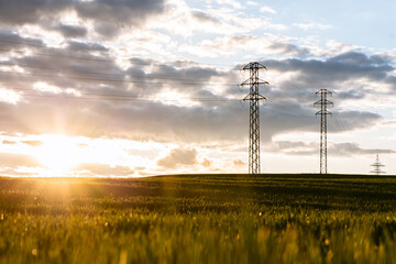 Electricity pylons in countryside at sunset with warm golden sunlight over green field and dramatic clouds, capturing contrast between nature and energy infrastructure