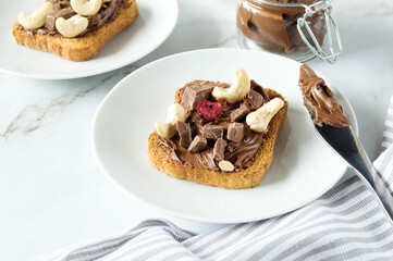 Spreading sweet chocolate cream, cashew, chocolate chunks onto toast on table.